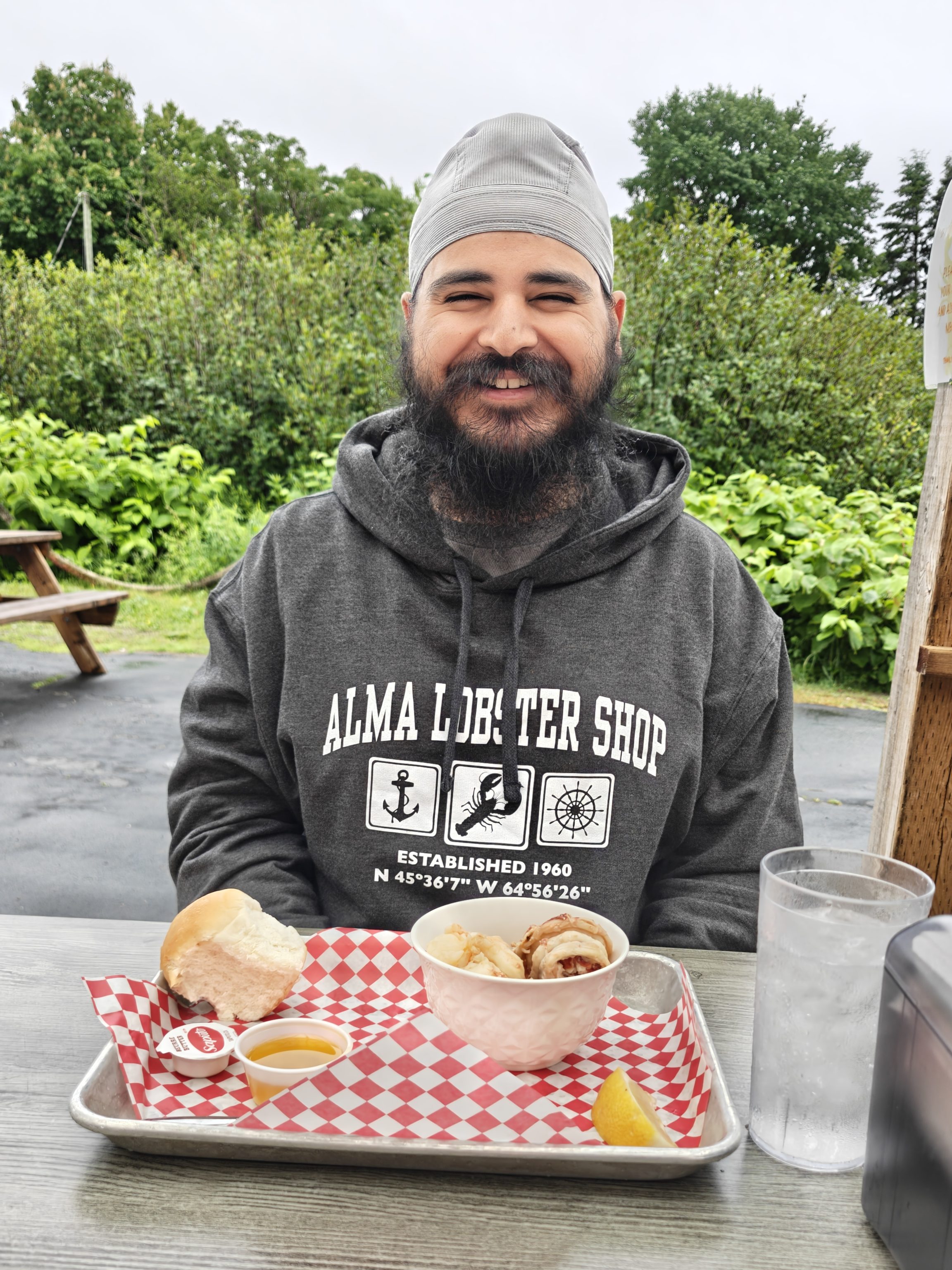 A smiling man wearing a grey sweatshirt and head covering sits at a table outdoors, enjoying a seafood meal with dishes presented on a red and white checkered tray, surrounded by greenery.